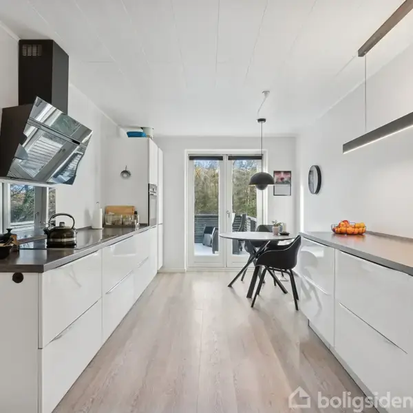 Et moderne køkken med hvide skabe strækker sig langs to vægge. Spisebordet med sorte stole står foran en glasdør, der fører ud til en terrasse. En frugtskål pynter på bordet.