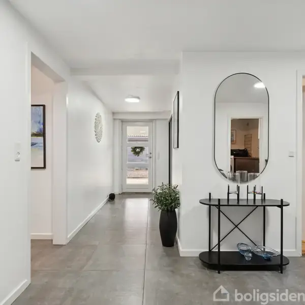 Simpel og moderne gang med en oval spejl og sort konsolbord til højre. Vægge er hvide, gulvet er gråt, og der er planter i en vase. En dør ses bagerst.