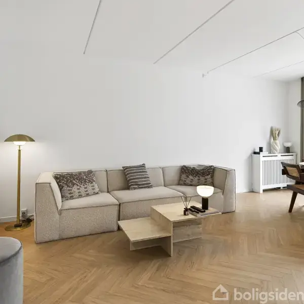 Sofa med puder står i en stue med trægulv, flankeret af gulvlampe og kaffebord. Til højre ses et spisebord med stole og store vinduer med gardiner.