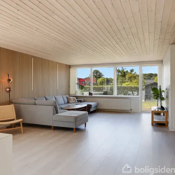 Stue med en stor, grå hjørnesofa og en træstol. Vinduerne slipper meget lys ind og giver udsigt til en have. Væggene har træpaneler, og gulvet er lyst.