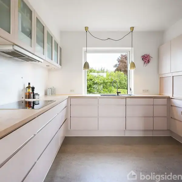 Et moderne køkken med lyse træskabe og en stor bordplade. En vase med gule blomster står på bordet. Vindue i midten giver udsigt til grønne omgivelser.