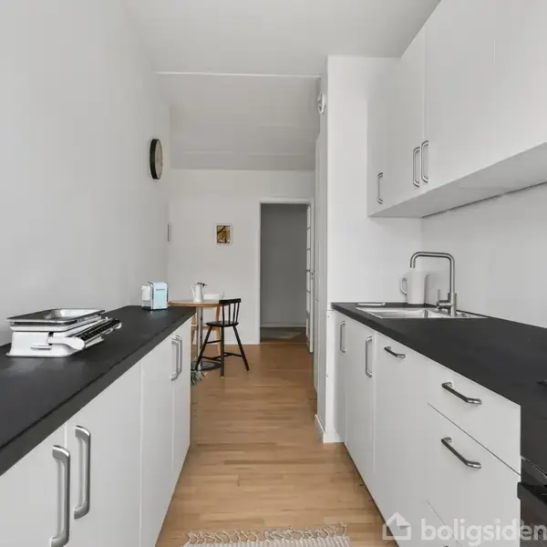 Moderne køkken med hvide skabe og sorte bordplader, ingen madlavning i gang. Lyse trægulve strækker sig til et spiseområde med bord og stol i baggrunden. Ingen tekst i billedet.