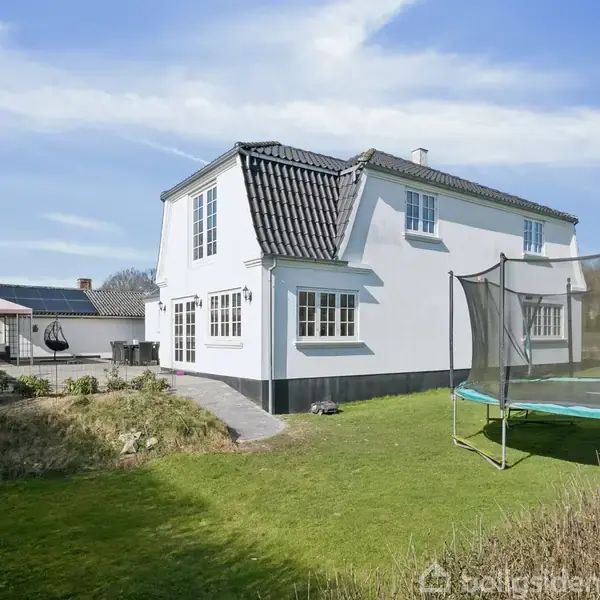 Hvidt to-etagers hus står stille i en grøn have med græs. Ved siden af huset er en trampolin, og i baggrunden ses et andet hus. Det er solrigt vejr.