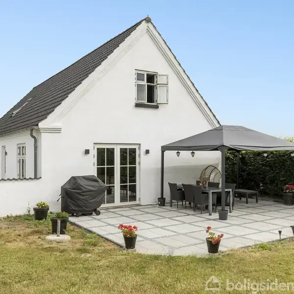 Hvidt hus med sort tag har en terrasse udstyret med en grill og et bord under en pavillon. Græsplæne med blomsterkrukker omgiver terrassen.