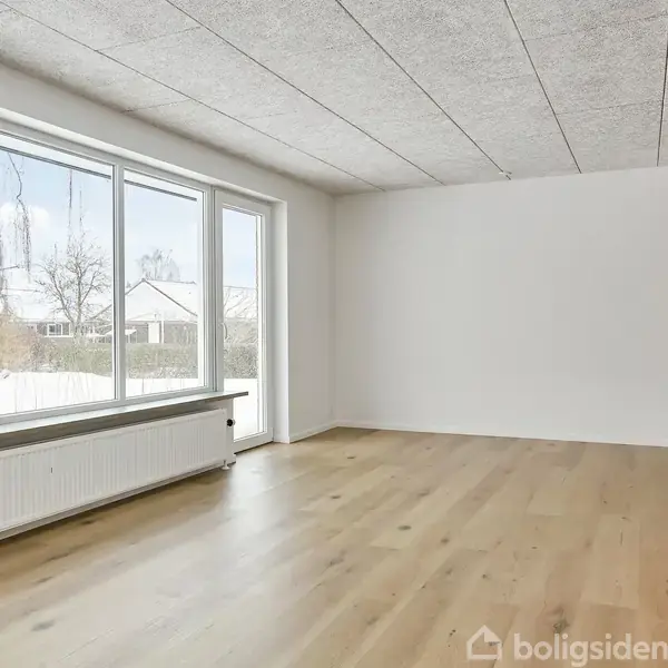 Rummelig stue med store vinduer, der slipper masser af lys ind, og parketgulv. I baggrunden ses snedækkede træer og huse gennem vinduerne. Rummet er lyst og indbydende.