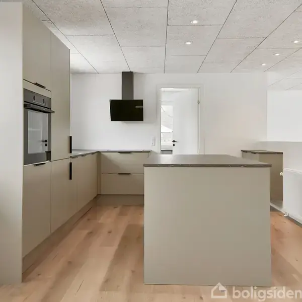 Et moderne køkken med lysegrå skabe og en central køkkenø. Rummet har trægulv og hvide vægge. En dør til et andet rum er åben, og der er en indbygget ovn.