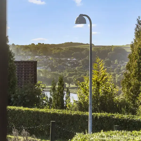 En gadelampe står stille i en frodig have med udsigt over en dal. Baggrundslandskabet består af grønne bakker og en nedgående sol, der skaber en lys, fredelig atmosfære.