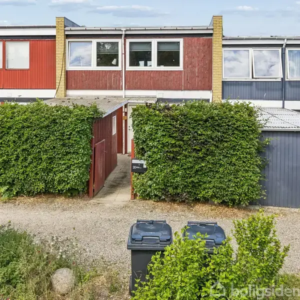 Rækkehus med røde og blå facader, står bag grønne hække. To skraldespande er placeret på en grusvej foran huset. Nabo huse ses til højre og venstre.