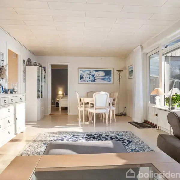 En stue har en lys indretning med hvidt møblement, herunder et rundt spisebord og stole. Store vinduer på højre side lader naturligt lys strømme ind. En sofa er delvist synlig.