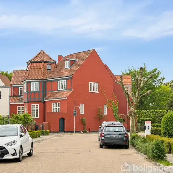 En rød murstensbygning med hvide vinduer står centralt, omgivet af grønne hække og træer. To biler er parkeret på vejen foran huset, under en blå himmel.