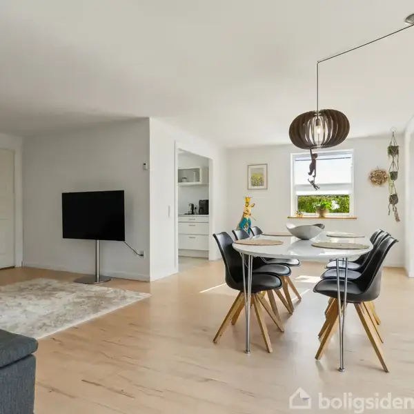 Et moderne spisebord med fire stole står i et lyst rum med trægulve. En lille stue med sofa og tæppe ses til venstre, og store vinduer lader lys strømme ind.