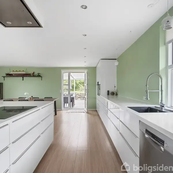 Hvidt, moderne køkken med stor køkkenø og indbygget kogeplade i et åbent rum. Grønne vægge og store vinduer skaber en lys atmosfære. Dobbelt glasdøre leder ud til haven.