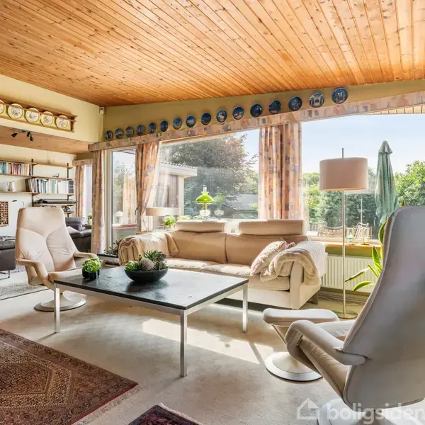 Stue med hvide lænestole og sofa omkring et rektangulært sofabord, overdækket med planter. Store vinduer bagved viser en terrasse og have. Loftet er træpanelet, og tallerkener pryder toppen af væggen.