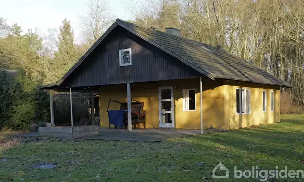 Et lille, gult hus står stille i en skovklædt have med græs og træer omkring. En terrassestol og et lille bord står under husets overdækkede veranda.