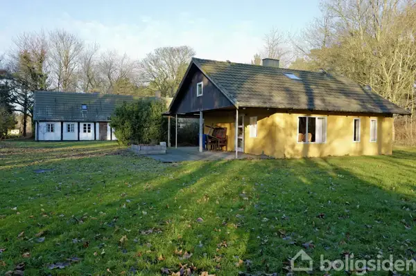 To huse står på en grøn græsplæne, omkranset af træer. Det forreste hus er gult med terrasse og havemøbler; det bageste hus er hvidt og halvt skjult af buske.
