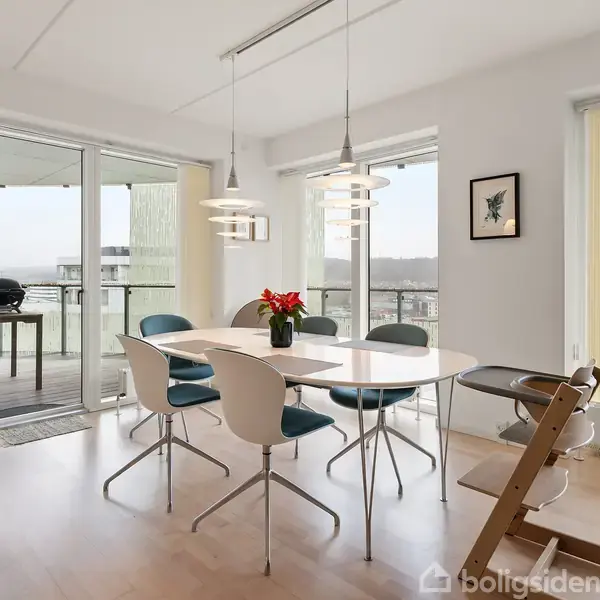 Et spisebord står i et veloplyst rum omgivet af moderne stole. En vase med røde blomster pryder bordet. Store vinduer åbner ud mod en balkon med udsigt over byen.