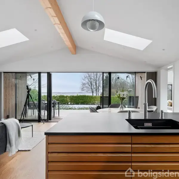 Køkkenø med vask står centralt i et moderne rum. Store glasdøre i baggrunden åbner udsigt til en terrasse og have. Lysindfald gennem loftsvinduer skaber en lys atmosfære.