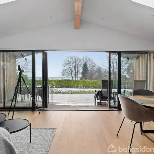 Stue med et spisebord og stole står foran en stor glasdør. En kikkert er placeret ved døren, der åbner ud til en terrasse med udsigt over en have.