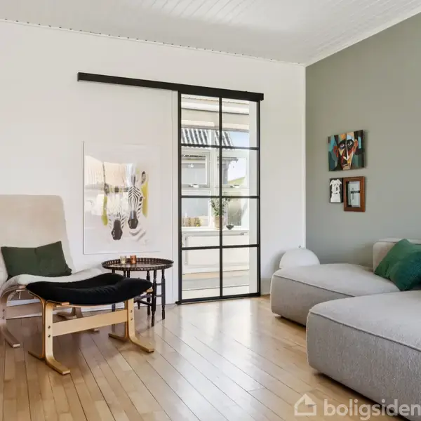 Stue med moderne indretning; en lys lænestol med skammel står ved siden af en sofa med farverige puder. En glasdør åbner mod udendørsområde; vægge prydes af kunstværker.