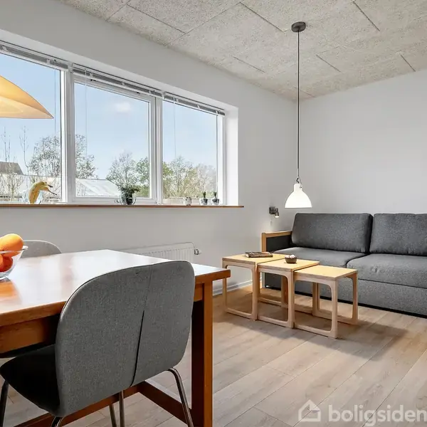 Gråt sofaarrangement ved siden af træbord med frugtskål, placeret under en pendellampe. Store vinduer bagved viser træer udenfor, mens træmøbler og lyse vægge skaber en moderne atmosfære.