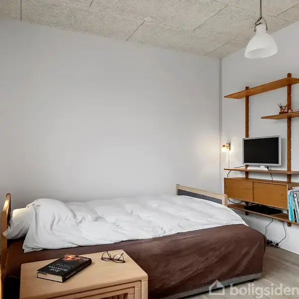 En seng med hvidt sengetøj står i et rum med en væghylde, hvor en lille skærm og bøger er placeret. En natlampe og planter dekorerer rummet.