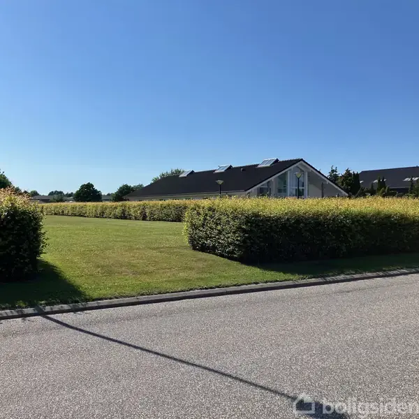 Rækkehuse ses i baggrunden omgivet af velplejede hække. En asfalteret vej strækker sig forbi, mens en lys lygtepæl står ved vejkanten under en klar blå himmel.