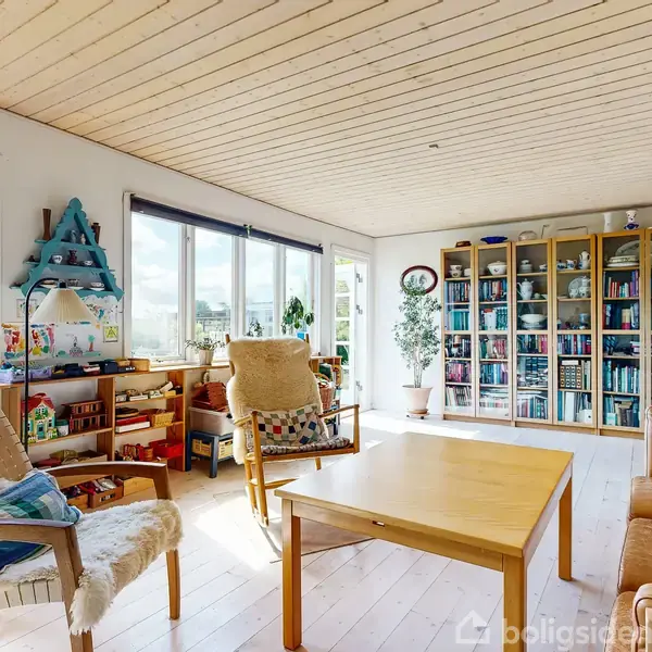 Stue med en sofagruppe og to lænestole omkring et træbord. Store vinduer lader lys ind. En reol fyldt med bøger og dekorationer står i hjørnet.