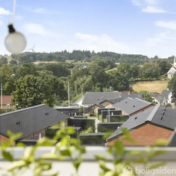Lyspære i en vinduesramme over en grøn udsigt; huse med sorte tage, træer og en kirke i baggrunden under en blå himmel med skyer.