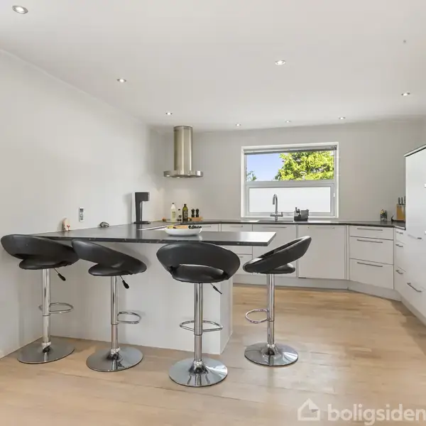 Køkken med hvide skabe og en mørk bordplade har fire sorte barstole ved køkkenøen. Et vindue over vasken slipper lys ind, og der er indbygget ovn til højre.