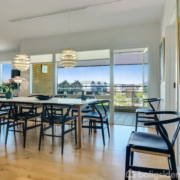 Spisebord med sorte stole står i et lyst rum nær store vinduer. Baggrunden viser en balkon med udsigt over hustage og det fri. To moderne lamper hænger over bordet.