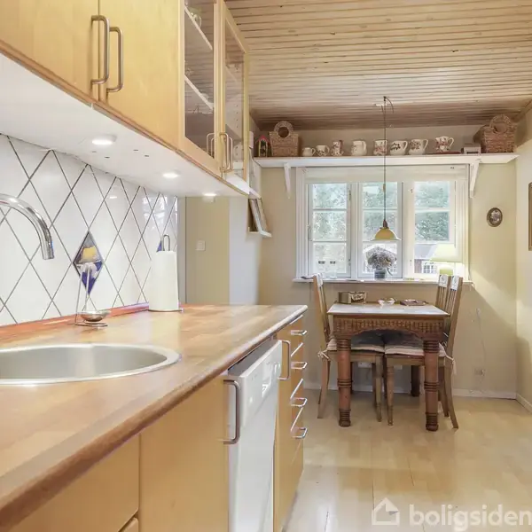 Køkken med træbordplade og vask, stilleben. Vægbeklædning med hvide fliser og blå detaljer. I baggrunden spiseområde med træbord, bænk, stole og vinduer omrammet af dekorationer.