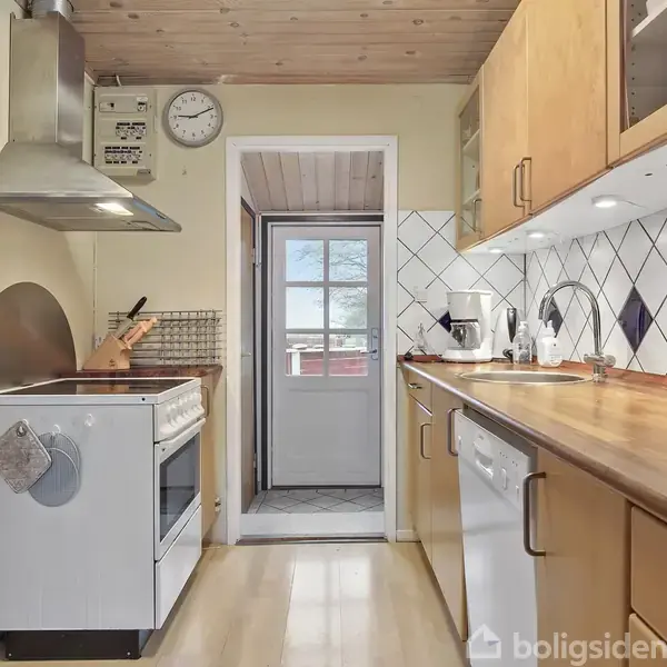 Et køkken med træloft og lyse træskabe. Der er en komfur med emhætte til venstre og en opvaskemaskine samt vask til højre. For enden ses en dør med vindue.