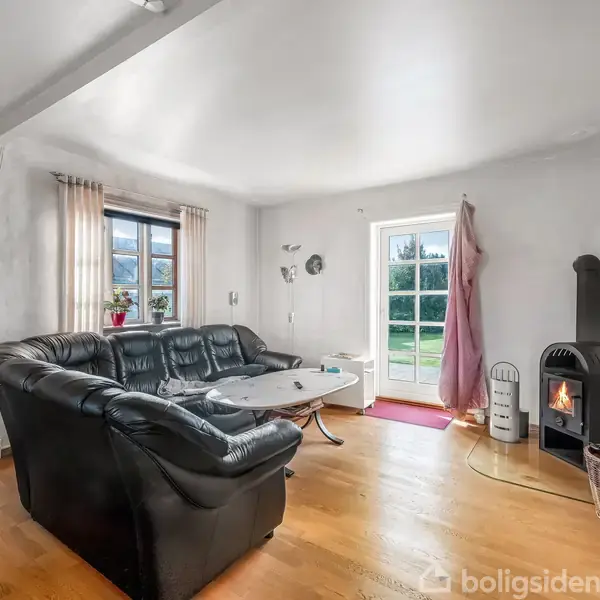 Sort hjørnesofa står ved sofabord i stue. Pejs brænder ved siden af åbent glasdør. Væghylde med bøger og billeder. Lyse gardiner dækker vindue. Trægulv dominerer rummet.