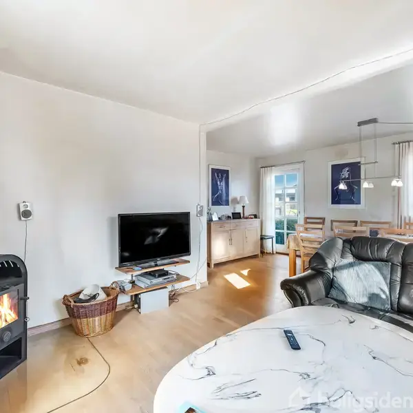 Brændeovn opvarmer stue med lædersofa og marmorbord. TV ved væggen, kurv til brænde ved siden. Spisebord og lysegardiner i baggrunden. Væglamper og dekorationer ses.