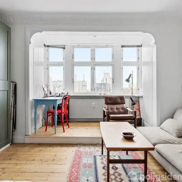 En stue med en beige sofa og et træbord. Ved vinduet i baggrunden står en rød stol foran et skrivebord. Et stort, grønt skab står til venstre.