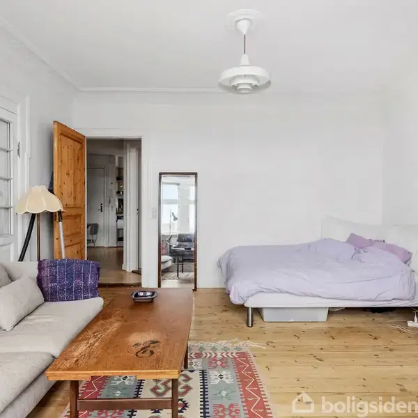 Gråt sofaarrangement med træbord og farverigt tæppe. Seng i baggrunden ved siden af et højt, grønt skab. Lyse vægge og trægulv i rummet.