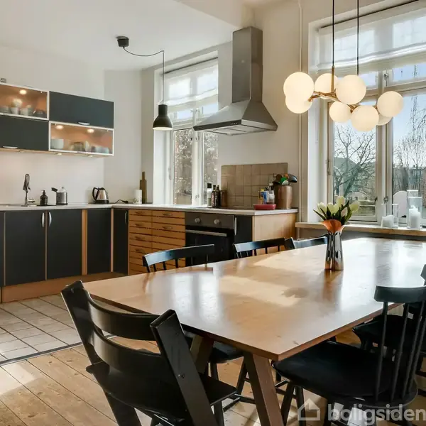 Køkkenbord med sorte stole står i et lyst og moderne køkken. Over bordet hænger en lampe, og i baggrunden ses køkkenskabe, vask og vinduer med udsigt til træer.