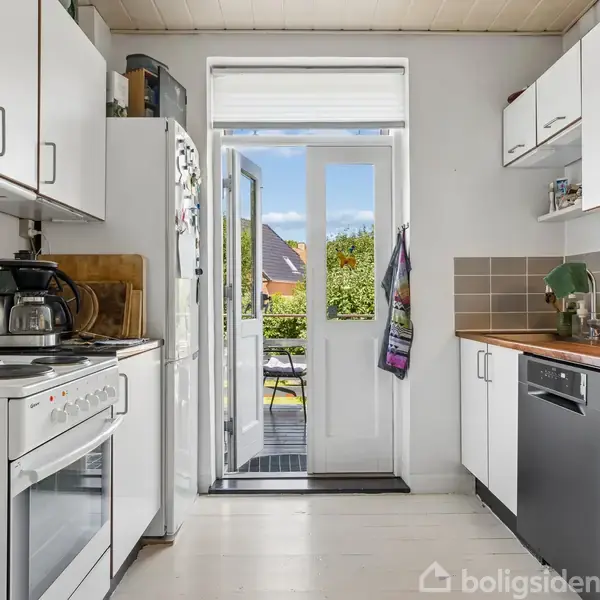 Stue med træbord, omgivet af stole, og en lænestol ved siden af reol. En lampe hænger over bordet. Åben dør fører til anden stue med sofa og store vinduer.