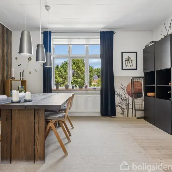 Et spisebord i træ med stole omkring står i et lyst rum. Der hænger tre lamper over bordet. Vindue i baggrunden med blå gardiner. Mørk reol til højre.