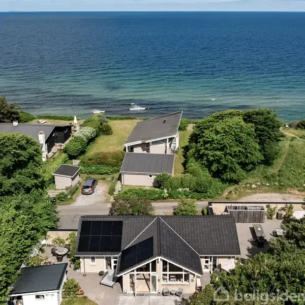 Huset med solpaneler på taget står stille i naturskønne omgivelser, tæt på havet. Omkringliggende hække og træer skaber en grøn ramme. Flere huse ses langs kysten.