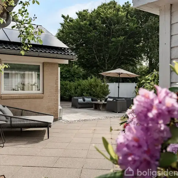 En gårdhave med fliser, hvor en loungesofa står foran et hus. I baggrunden ses et havebord med parasol. Blomsterbusk i forgrunden og træer omringer området.
