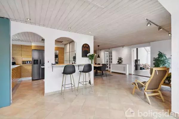 Køkkenalrum med køkkenbar og to barstole, åbner ind til spisestue med trægulv og moderne møbler, omgivet af hvidmalede vægge og lofter.