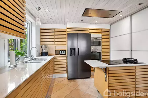 Et moderne køkken med trægulv og træpaneler på væggene. Et sort køleskab står mellem en ovn og et komfur med emhætte. En stor vask er under et vindue.