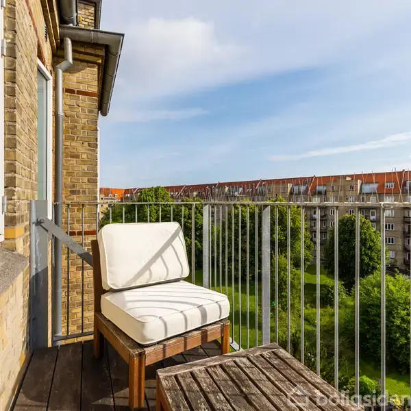 En balkon med en hvid polstret stol og træbord set mod en lejlighedskompleksets have. Omkranset af metalgelænder og murstensbyggeri i baggrunden. Klar, blå himmel ovenover.