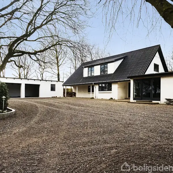 Hvidt hus med sort tag står stille på en grusbelagt gårdsplads, omgivet af træer. Ved siden af huset ses en garagebygning.