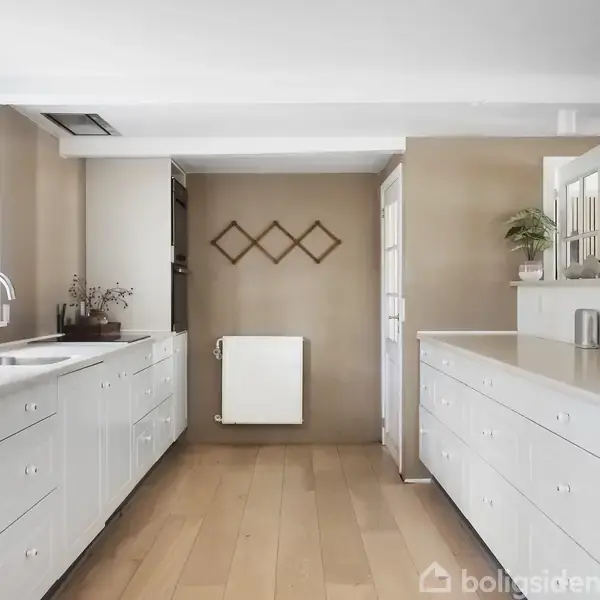 Køkken med hvide skabe og træbordplader, stålvask og arrangement af planter. Rummet har lyse trægulve og beige vægge, og en radiator ses på væggen under simpelt vægkunst.