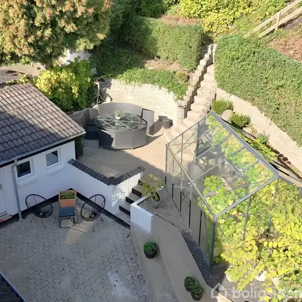 En brostensbelagt gård med et lille hvidt hus, omgivet af grønne planter. Et drivhus står til højre, og et rundt havebord sæt er placeret på en øvre terrasse.