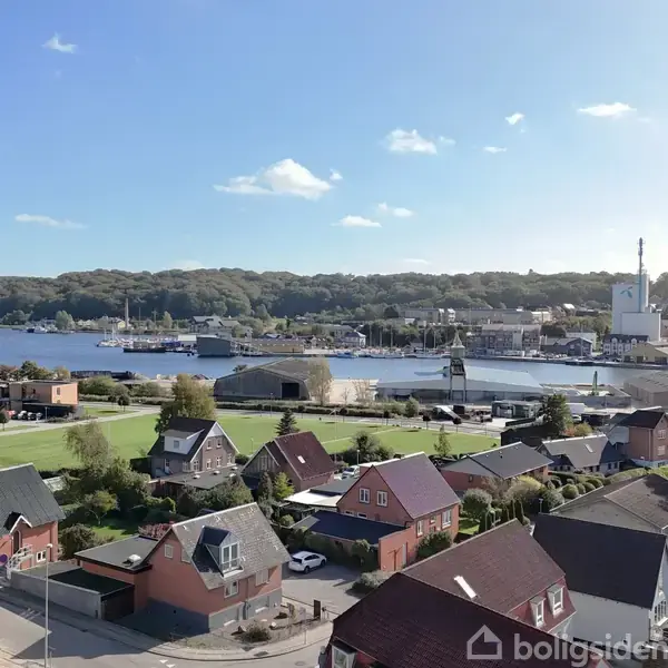 Røde og grå huse ligger i en række med udsigt over en rolig bugt, hvor både ligger ved kajen. Skoven omkranser horisonten under en klar, blå himmel.