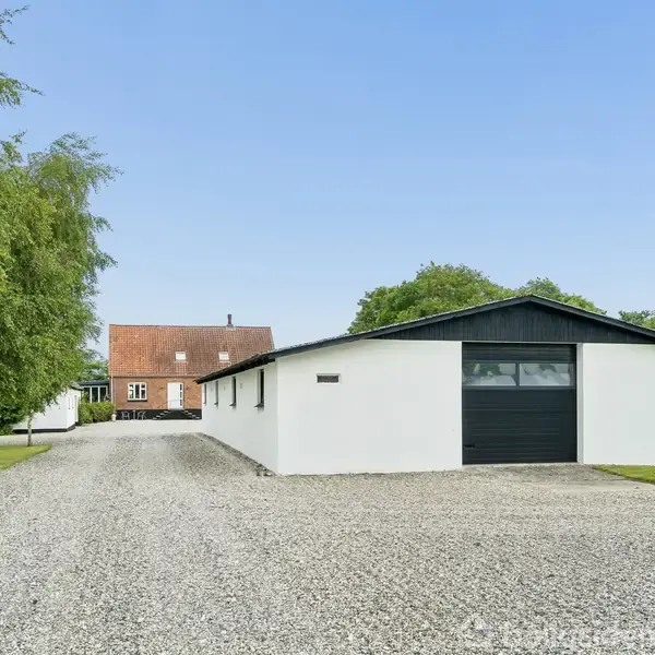 Et hvidt lagerbygning med sort tag står ved siden af en grusvej. I baggrunden ses et murstenshus med rødt tag. Græsarealer og træer findes omkring bygningerne.