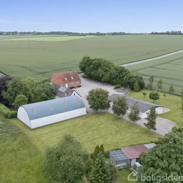 Gårdkompleks med flere bygninger, herunder et hus med rødt tag, omgivet af marker og træer. En lang grusvej forbinder ejendommen til den omkringliggende landlige landskab.
